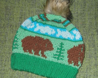 May include: Green knitted hat with a brown bear design, blue and white stripes, and a brown faux fur pom pom.