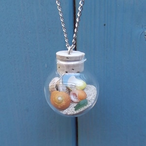 May include: A silver chain necklace with a small glass bottle pendant filled with sand, seashells, and a small piece of sea glass.