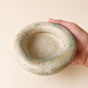 May include: A round, shallow ceramic bowl with a thick, rounded rim. The surface features a speckled beige base with mottled green and brown glaze accents distributed unevenly around the exterior and the inner edge of the rim.
