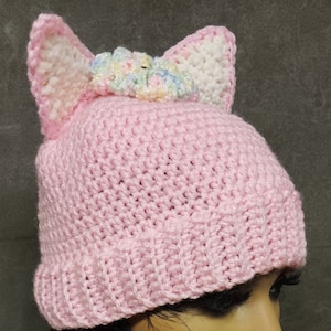 May include: Pink crocheted beanie with white cat ears and a colourful flower embellishment on top.