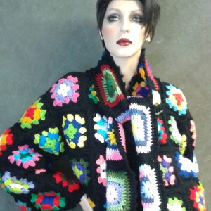May include: A black crochet cardigan with a colorful granny square pattern. The cardigan has a shawl collar and is open in the front.