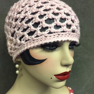 May include: A pink crocheted hat with a delicate openwork design. The hat is worn by a mannequin with dark hair and red lipstick.