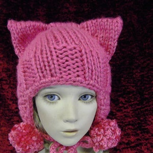 May include: A pink knitted hat with cat ears and pom-poms. The hat is worn by a doll with blonde braids.