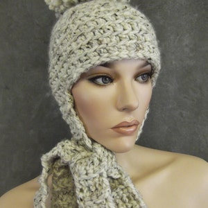 May include: A hand-knitted, cream-colored hat with ear flaps and a matching scarf. The hat features a large pom-pom on top. The yarn has flecks of black and brown. The scarf is designed to be tied around the neck.