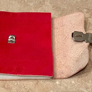 May include: A red leather journal with a metal clasp. The journal is open, revealing the blank pages inside. The cover has a textured design. The journal is a classic style, ideal for writing or sketching.