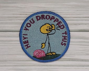 May include: Embroidered patch with a blue border and a light blue background. The patch features a yellow stick figure with a surprised expression, reaching towards a pink brain. Text around the border reads "HEY! YOU DROPPED THIS."