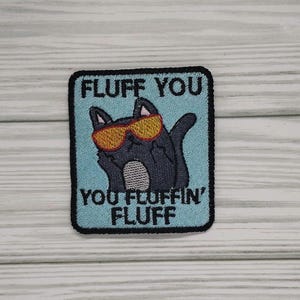 May include: Embroidered patch featuring a black cat wearing orange sunglasses, with the text "FLUFF YOU YOU FLUFFIN' FLUFF" on a light blue background. The patch has a black border and is likely for clothing or accessories.