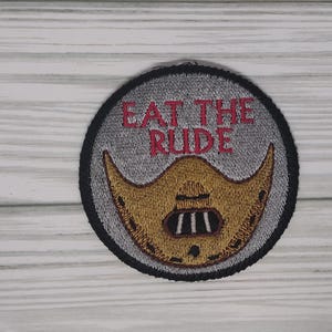 May include: A round, gray and black embroidered patch with the text "Eat the Rude" in red lettering and a yellow, stitched image of a face mask.