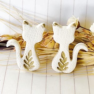 May include: Two white cat-shaped ornaments with gold accents on the ears and a gold leaf design on the body. The ornaments are set against a backdrop of dried wheat and a striped surface.