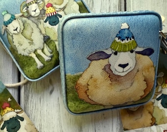 May include: Square, blue coin purses with sheep illustrations wearing colorful knit hats. The purses have a textured appearance and are set against a light wood background. Tags with the name "Emma Balle" are also visible.