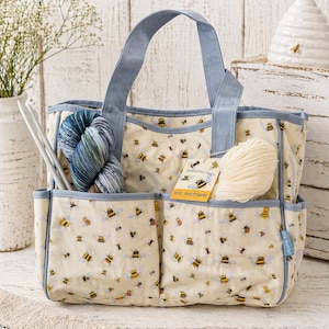 May include: Cream-colored tote bag with a bee pattern and light blue trim. The bag has two exterior pockets and a pair of handles. Inside are knitting needles and yarn. The text on the tag reads "Beautiful bees..." and "The Heyman's".