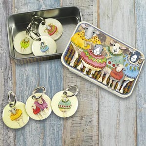May include: A collection of sheep-themed keychains and a decorative box. The keychains feature sheep illustrations wearing colorful sweaters. The box has a rectangular shape and a raised relief design of sheep in sweaters. The items are on a wooden surface.