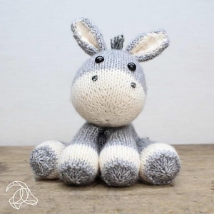 May include: A handmade knitted donkey plush toy in gray and white. The donkey has large ears, black button eyes, and a white muzzle. The plush is sitting with its legs outstretched.