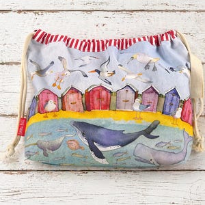 May include: Drawstring bag with a beach scene featuring colorful beach huts, seagulls, whales, and fish. The bag has a light blue background with a red and white striped top. The bag is made of fabric and has a drawstring closure.