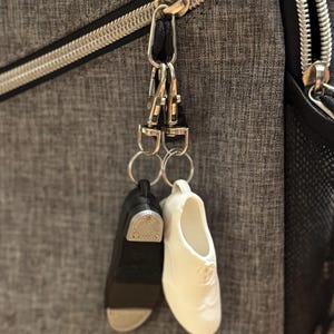 May include: A pair of tap shoe keychains, one black and one white, hanging from a silver clasp attached to a gray fabric bag. The black shoe has a silver tap plate. The white shoe has a sculpted design.