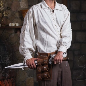 May include: A man wearing a white shirt with lace detailing and brown pants. He is holding a silver sword in his right hand and a brown leather belt with a pouch on his left hip.