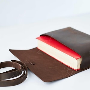 May include: A brown leather book cover with a red book inside. The cover has a strap that can be used to close it.