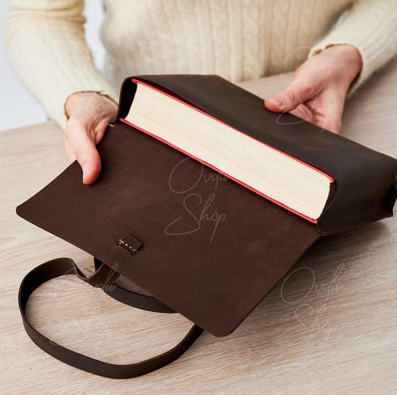 May include: A brown leather book cover with a strap. The cover is open to reveal a book with white pages and a red spine.
