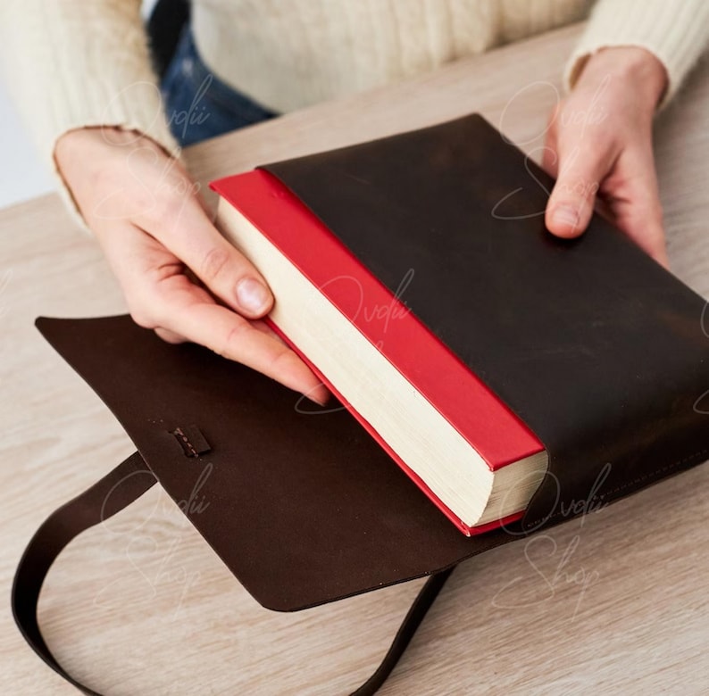 May include: A brown leather book cover with a red spine and a strap. The cover is open and a book is inside.