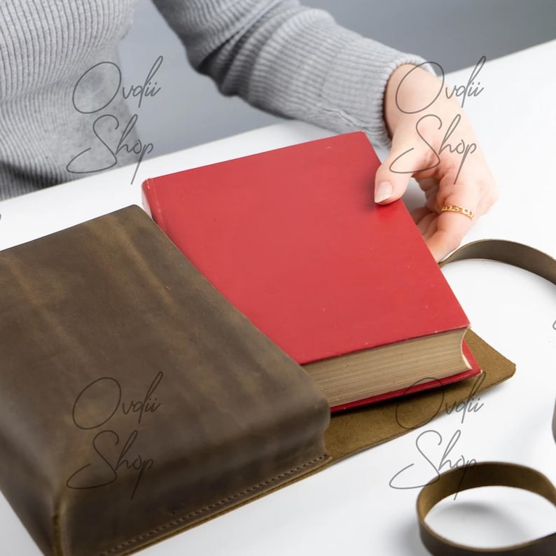 May include: A brown leather book sleeve with a red book inside. The sleeve has a flap that closes with a strap. The strap is made of the same leather as the sleeve. The sleeve is lying on a white surface.