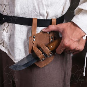 May include: A brown leather knife sheath with a metal buckle and a black leather belt. The sheath is attached to the belt and has a knife inside.