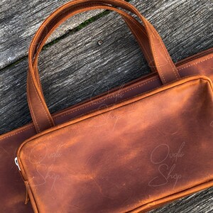 May include: A brown leather tote bag with a large flap and a zipper closure. The bag has a top handle and a shoulder strap. The bag is made of high-quality leather and has a distressed look.