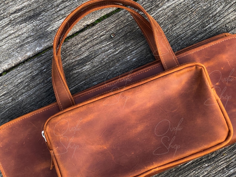 May include: A brown leather tote bag with a large flap and a zipper closure. The bag has a top handle and a shoulder strap. The bag is made of high-quality leather and has a distressed look.