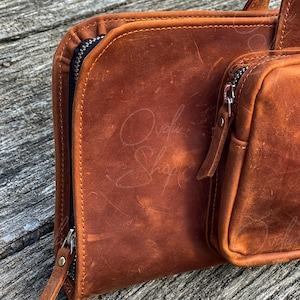 May include: A brown leather zippered pouch with a smaller zippered pouch attached. The pouches are made of distressed leather and have a rustic look.
