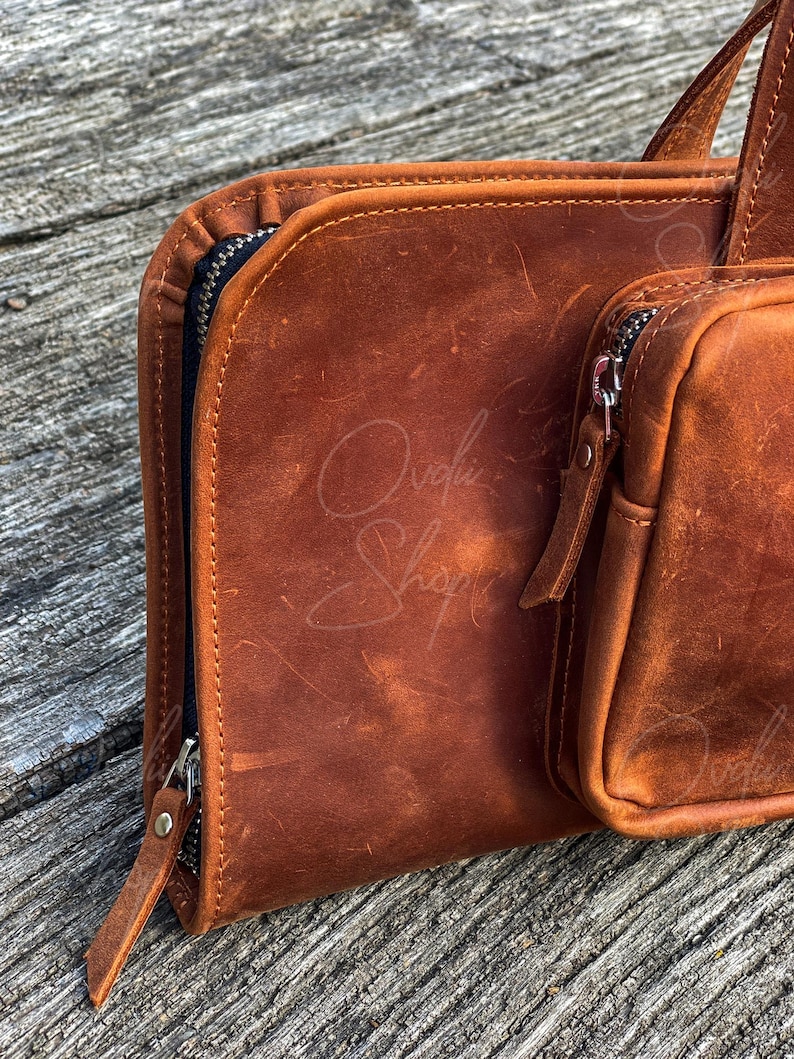 May include: A brown leather zippered pouch with a smaller zippered pouch attached. The pouches are made of distressed leather and have a rustic look.