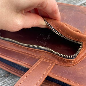 May include: A brown leather bag with a zippered pocket and a handle. The zipper is open and the inside of the bag is visible.