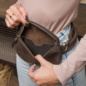 May include: A brown leather fanny pack with a zipper closure. The pack is worn around the waist with a brown leather belt. The interior of the pack is visible, revealing a pocket. The pack is made of a dark brown leather.