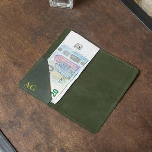 May include: A dark green leather wallet with a 20 Euro bill partially visible. The wallet has the initials "AG" in gold lettering on the front. The wallet is rectangular and appears to be made of suede.
