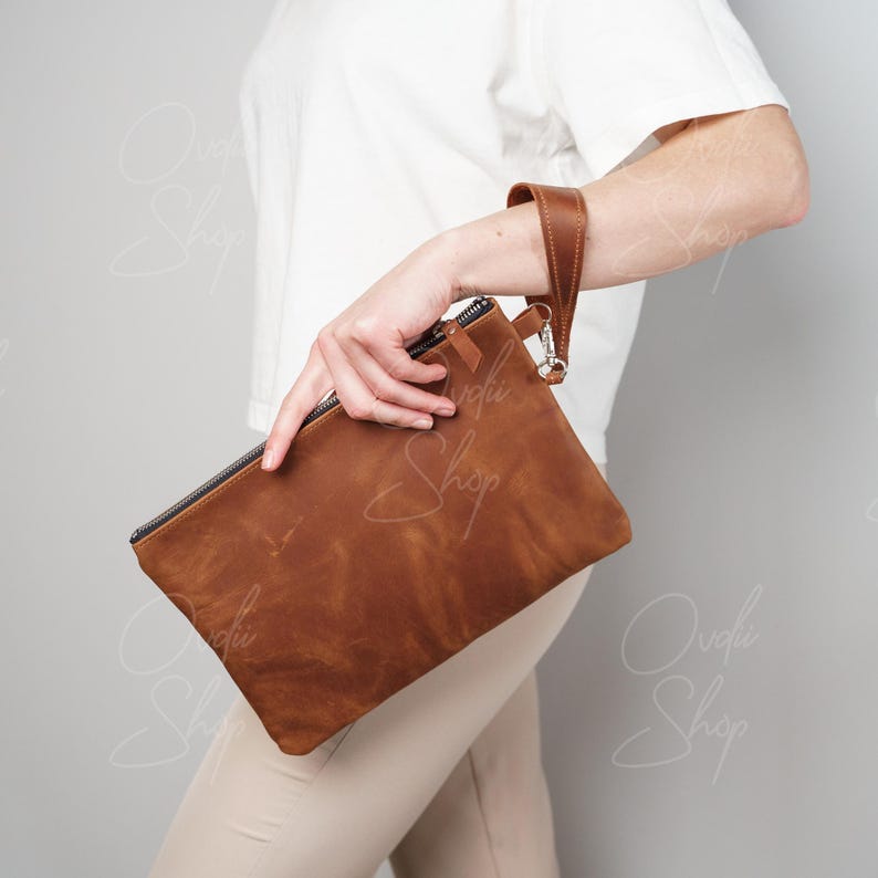 May include: A brown leather clutch with a wrist strap. The clutch has a zipper closure and a textured leather finish.