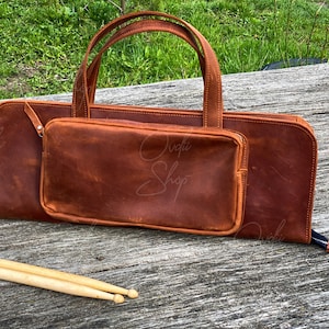 May include: A brown leather bag with a zippered pocket and a handle. The bag is sitting on a wooden surface with two wooden drumsticks in front of it.