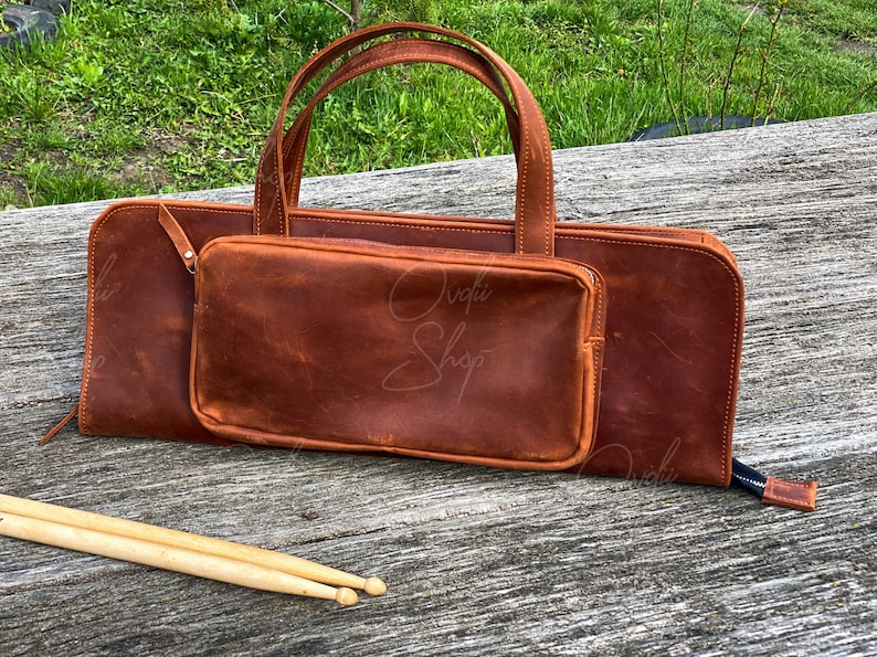 May include: A brown leather bag with a zippered pocket and a handle. The bag is sitting on a wooden surface with two wooden drumsticks in front of it.