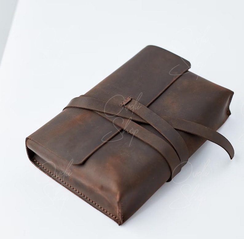 May include: A brown leather clutch with a strap that wraps around the front and ties at the top. The clutch has a distressed look and is made of high-quality leather.