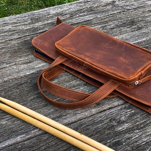 May include: A brown leather drumstick bag with a zipper closure and a separate compartment for drumsticks. The bag has a strap for carrying.