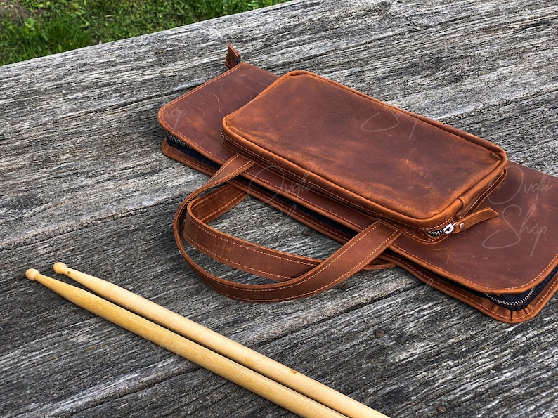 May include: A brown leather drumstick bag with a zipper closure and a separate compartment for drumsticks. The bag has a strap for carrying.