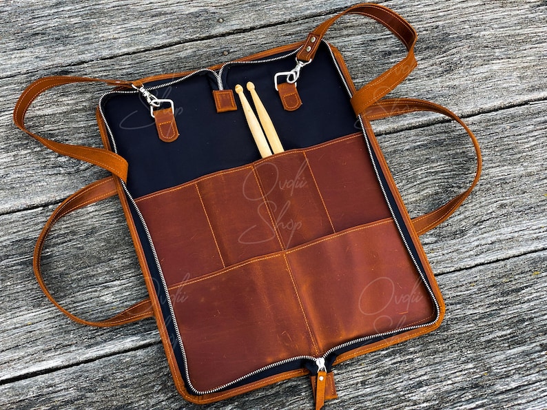 May include: A brown leather drumstick case with a zipper closure and multiple pockets. The case has two adjustable straps and is open to reveal two wooden drumsticks.