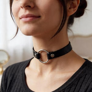 May include: A black leather choker necklace featuring a large silver ring. The choker is fastened with a buckle. This necklace is a fashionable accessory.