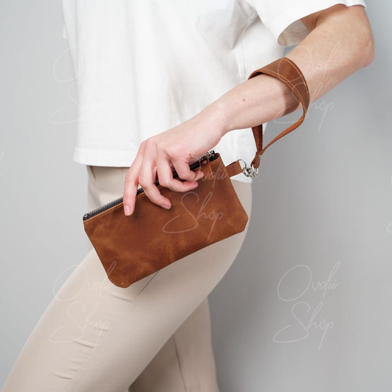 May include: A brown leather wristlet with a strap. The wristlet is held by a person wearing a white shirt and beige pants.