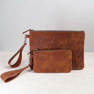 May include: Two brown leather zippered pouches with silver hardware and matching straps. The larger pouch is slightly wider than the smaller pouch.