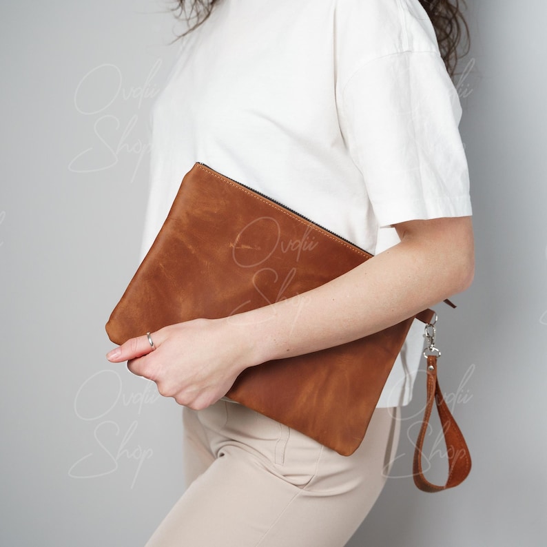 May include: A brown leather clutch with a silver zipper and a matching brown leather strap. The clutch is held by a person wearing a white shirt and beige pants.