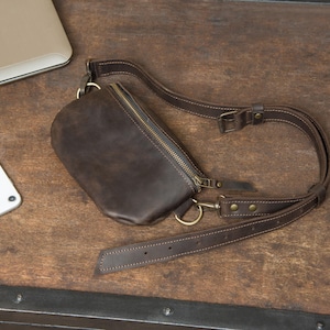 May include: A dark brown leather fanny pack with a zipper closure and adjustable strap. The bag has a rectangular shape with rounded edges and brass-colored hardware. The strap is also brown and has a buckle for adjusting the length.