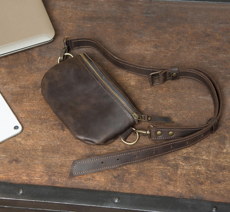 May include: A dark brown leather fanny pack with a zipper closure and adjustable strap. The bag has a rectangular shape with rounded edges and brass-colored hardware. The strap is also brown and has a buckle for adjusting the length.