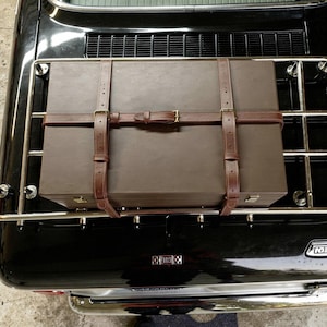 May include: A brown rectangular box secured to a car's luggage rack with brown leather straps. The box is a muted brown color, and the straps have brass buckles. The car is black.