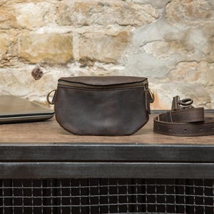 May include: A dark brown leather fanny pack with a zipper closure and gold-colored hardware. A matching leather belt is coiled next to the bag. The bag and belt sit on a wooden surface with a brick wall background.