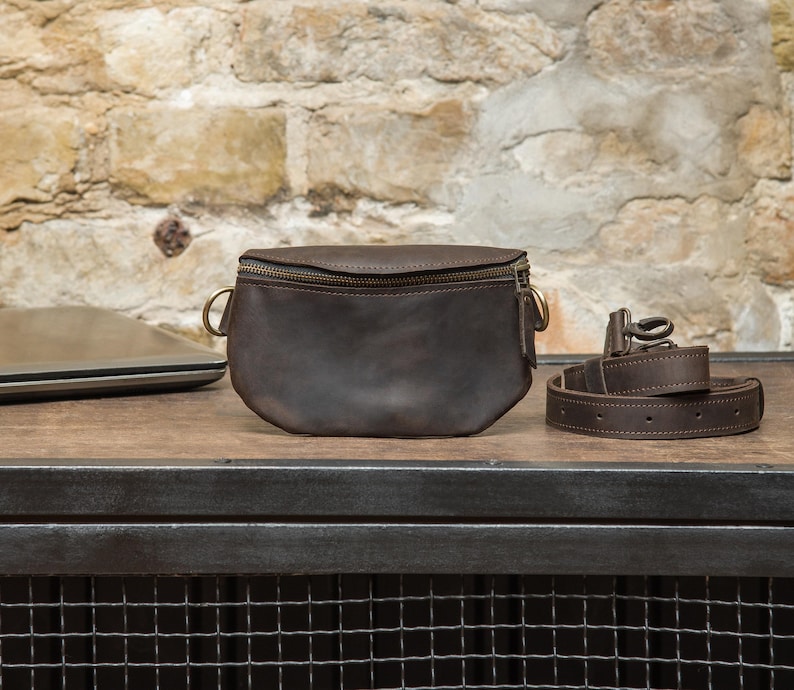 May include: A dark brown leather fanny pack with a zipper closure and gold-colored hardware. A matching leather belt is coiled next to the bag. The bag and belt sit on a wooden surface with a brick wall background.