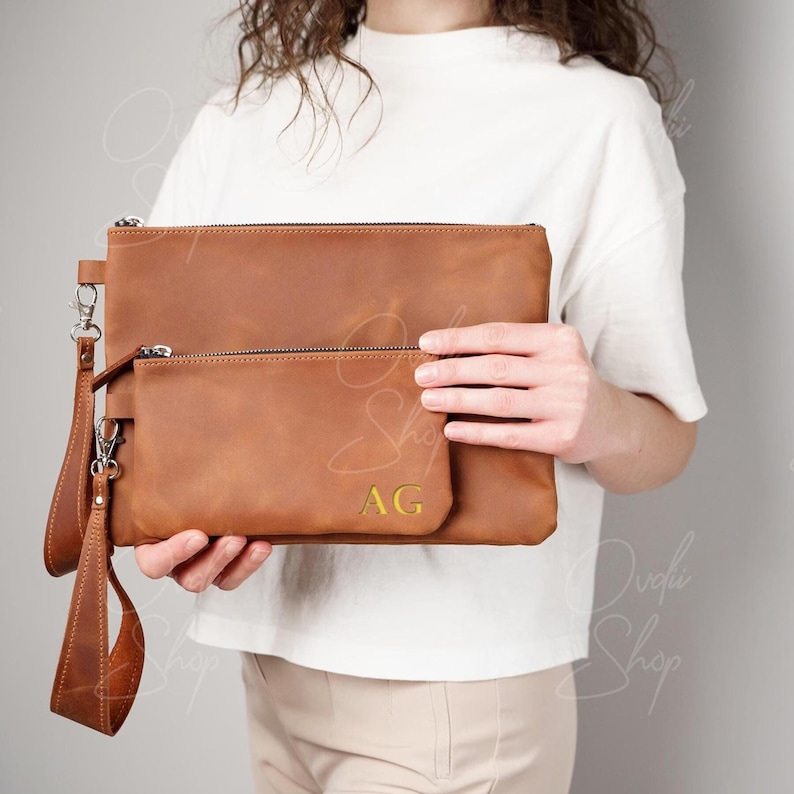 May include: A brown leather clutch with a smaller matching pouch. The clutch has a zipper closure and a wrist strap. The pouch has a zipper closure. The clutch has the initials "AG" embossed on the front.