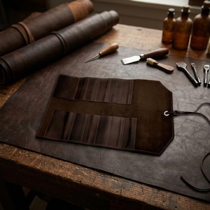 May include: A dark brown leather tool roll, open to show multiple pockets. Rolled leather sheets and various leatherworking tools are visible on a wooden work surface. The roll has a strap for securing it.